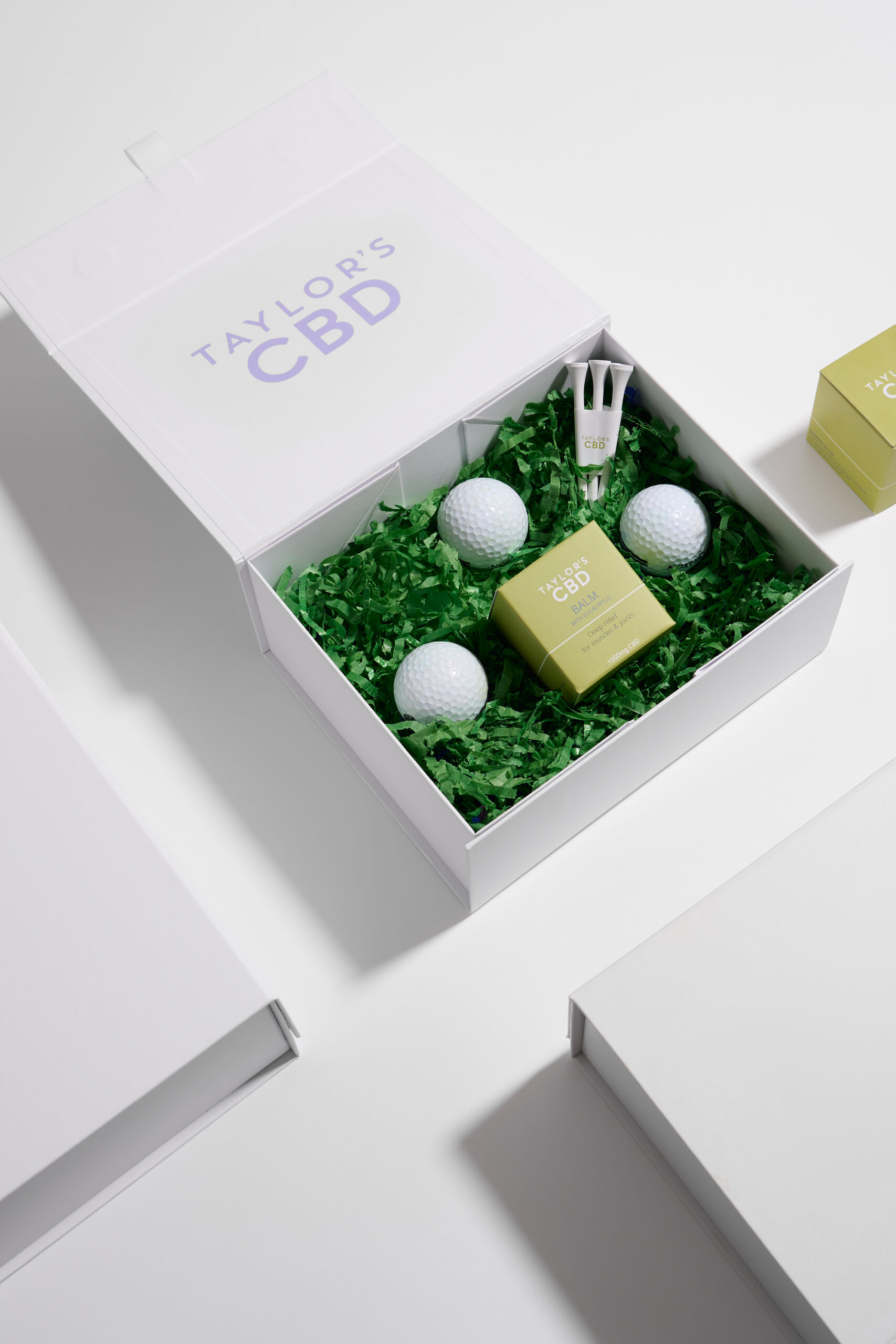 Overhead view of a white box containing balm and golf balls on a white background with strong shadows