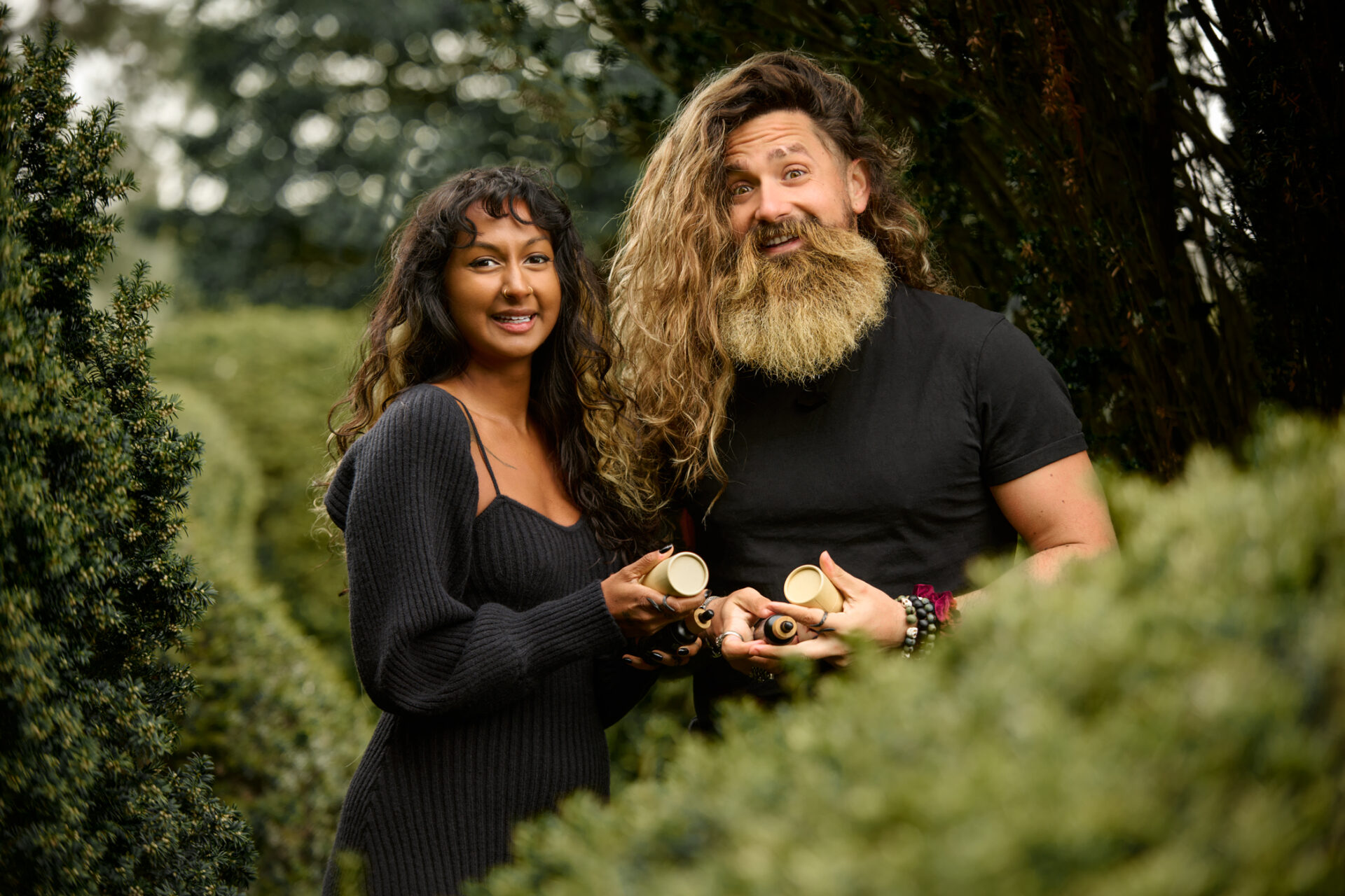 Couple headshot of a man and woman standing in the woods, smiling and holding their hair product, surrounded by natural greenery.