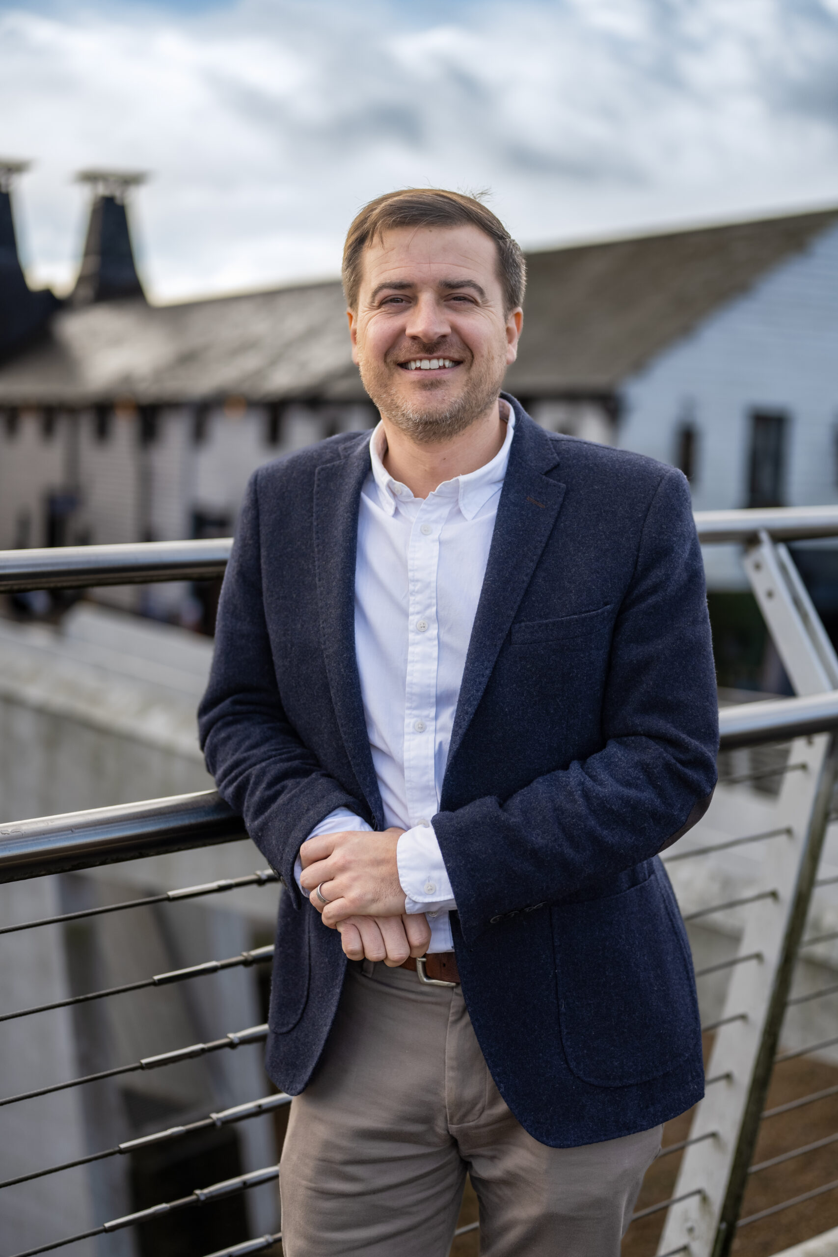 Outdoor headshot of a man standing on a bridge with buildings visible in the background.