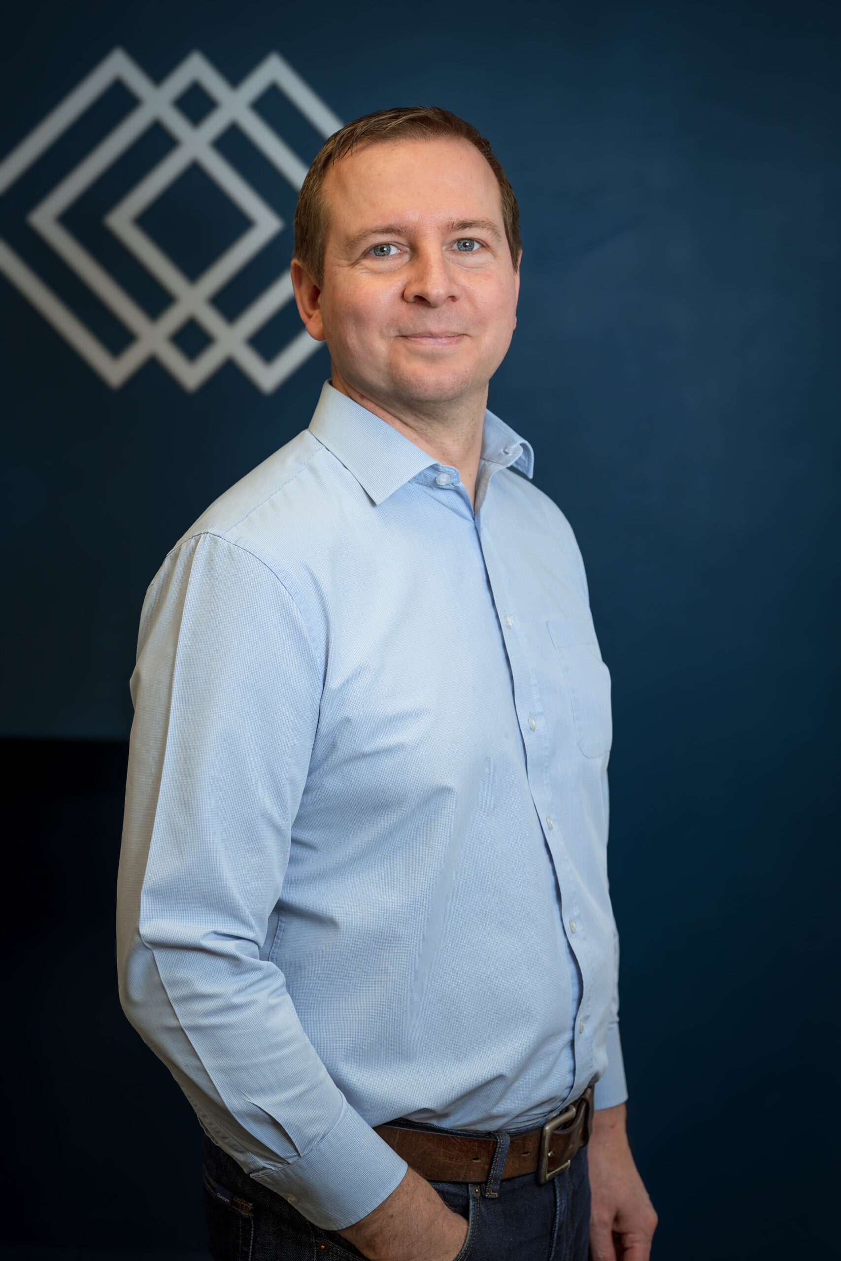 Professional headshot of a man standing in front of a dark blue wall with a company logo in the background, conveying confidence and brand identity.