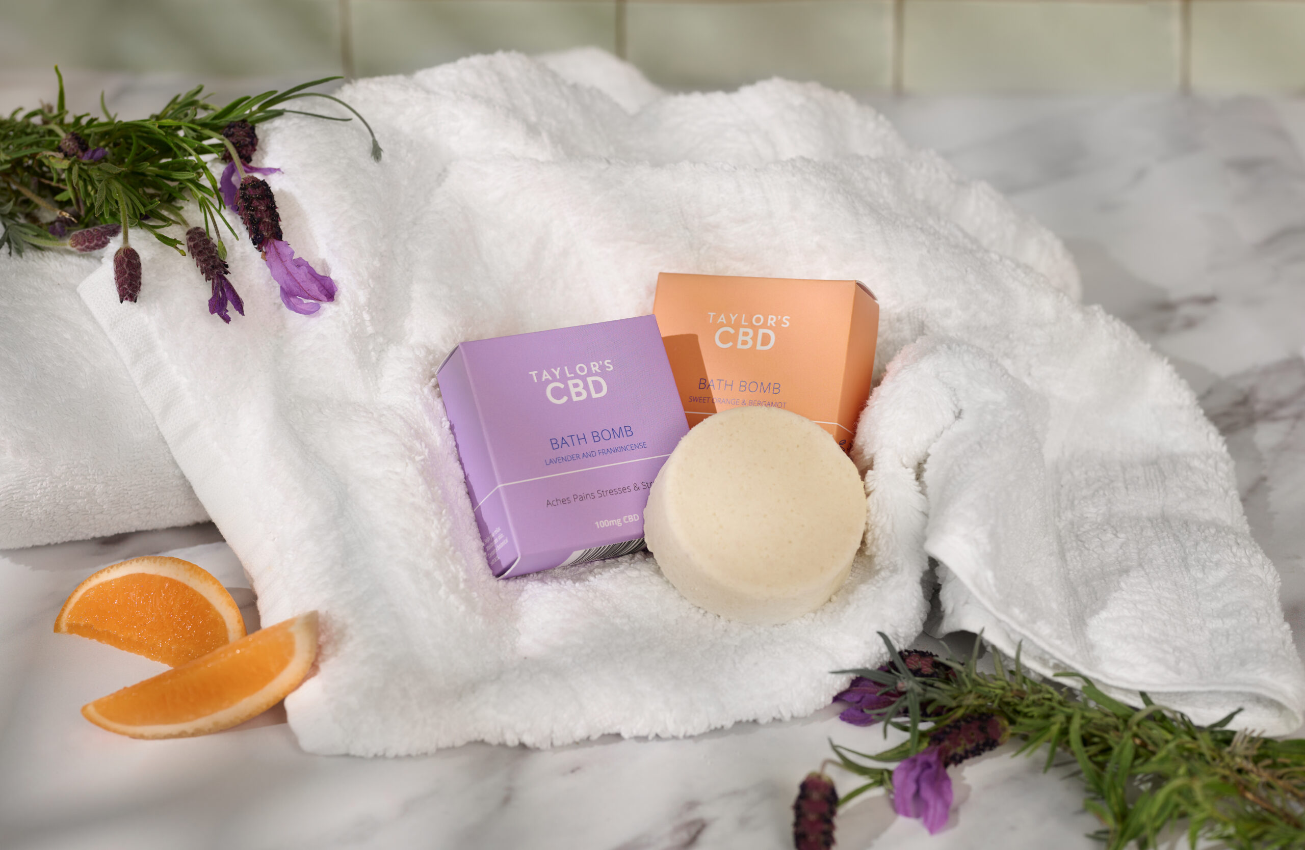 Two product boxes and a single bath bomb placed on a soft white towel in a bright bathroom setting, creating a clean and relaxing spa-like atmosphere.
